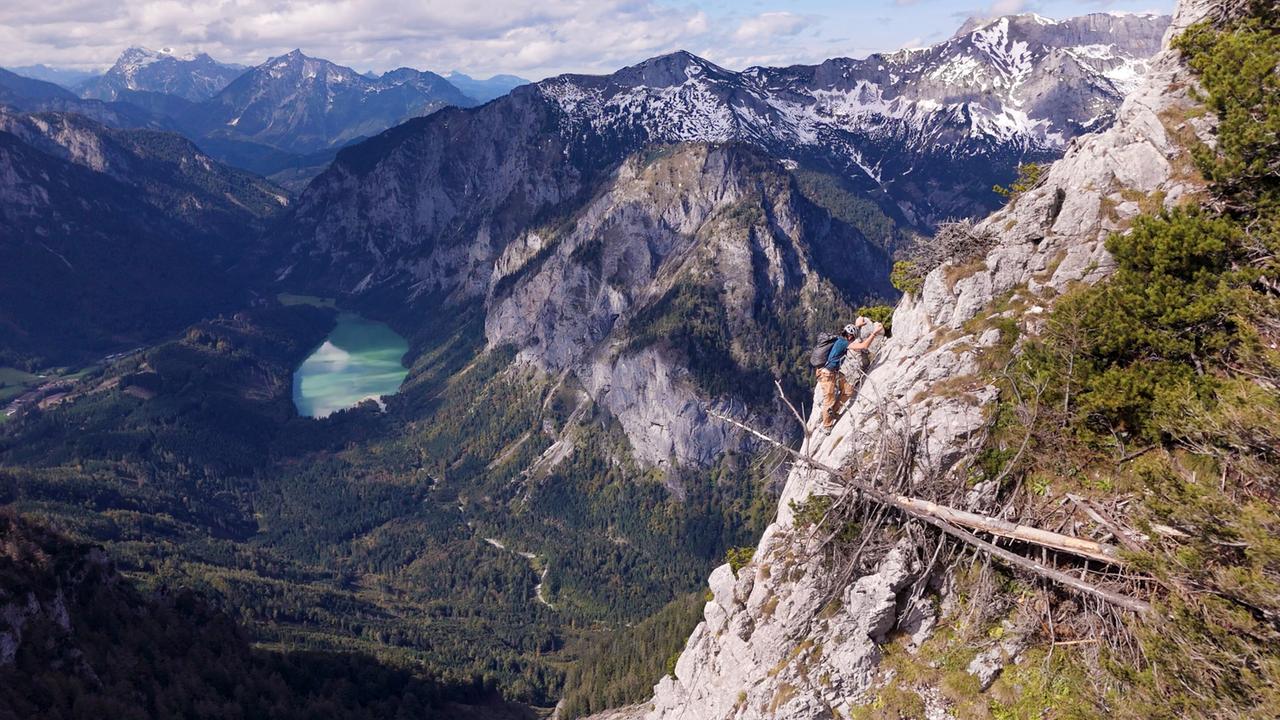 Bergwände mit einem Kletterer im Vordergrund, Berge und ein türkisfarbener Bergsee von oben gesehen