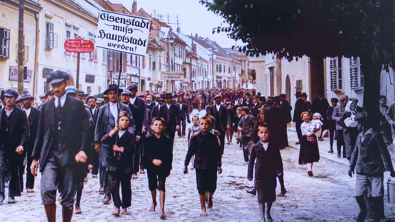 Das Bild zeigt eine historische Szene aus Eisenstadt, wahrscheinlich während einer Demonstration oder eines Festumzuges für den Status als Hauptstadt.   Im Vordergrund sind mehrere Männer und Kinder zu sehen, die barfuß und in traditionellen, eher einfachen Kleidern gekleidet sind. Die Kinder laufen fröhlich, während die Männer ernst blicken. Die Männer tragen Hüte, und einige halten Schilder in der Hand. Eines der Schilder trägt die Aufschrift "Eisenstadt muss Hauptstadt werden!"  Im Hintergrund ist eine Gruppe weiterer Menschen zu erkennen, die ebenfalls durch die Straße gehen. Die Umgebung zeigt typische, historische Gebäude mit Fenstern und Türen. Die Straße ist gepflastert, und die Luft scheint mild und klar zu sein. Bäume sind am Bildrand sichtbar, und das gesamte Szenario ist in lebhaften Farben gehalten, trotz der nostalgischen Ausstrahlung des historischen Kontexts.   Das Bild vermittelt ein Gefühl der Gemeinschaft und des Engagements für die Stadt Eisenstadt.