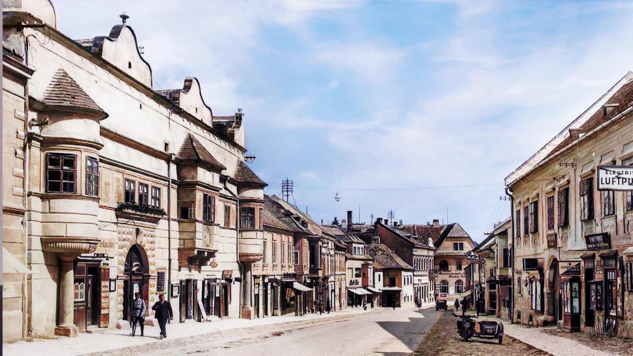 Das Bild zeigt die untere Hauptstraße in Eisenstadt, Österreich, zu einer historischen Zeit. Die Straße ist gepflastert und hat eine leicht abfallende Neigung. Auf beiden Seiten der Straße stehen historische Gebäude mit unterschiedlichen architektonischen Stilen.   Links befinden sich höher gebaute Gebäude, die aus hellem Stein und mit geschweiften Dachformen bestehen. Einige Fenster sind mit Holzläden versehen. Rechts erkennt man ebenfalls alte Häuser, die eine Mischung aus hellen und pastellfarbenen Fassaden haben. Geschäftssschilder sind sichtbar, darunter eines mit dem Wort "Luftpunkt".   Auf der Straße sind zwei Personen zu sehen, die aneinander vorbeigehen, sowie ein altes Automobil, das parkt. Der Himmel ist teilweise bewölkt mit einigen blauen Stellen. Der Gesamteindruck vermittelt eine ruhige, historische Atmosphäre, die an vergangene Zeiten erinnert.   Das Bild spielt auf die Bedeutung von Eisenstadt als kulturelles und politisches Zentrum des Burgenlandes an und erinnert an die 100-jährige Geschichte der Stadt als Landeshauptstadt.
