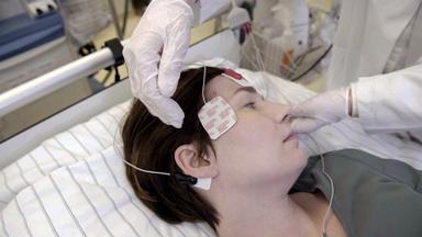 Frau liegt in einem Krankenhausbett, an ihrem Kopf sind Elektroden befestigt.