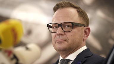 Ein Mann mir schwarzer Brille und Anzug schaut über die linke Schulter in die Kamera.