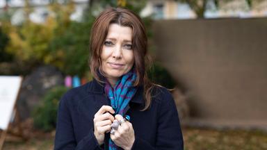 Porträtfoto einer Frau mit langen braunen, nach hinten gebundenen Haaren. Sie trägt einen dunkelblauen Mantel mit einem blau-lila-gemusterten Schal und viele große Silberringe an der linken Hand.