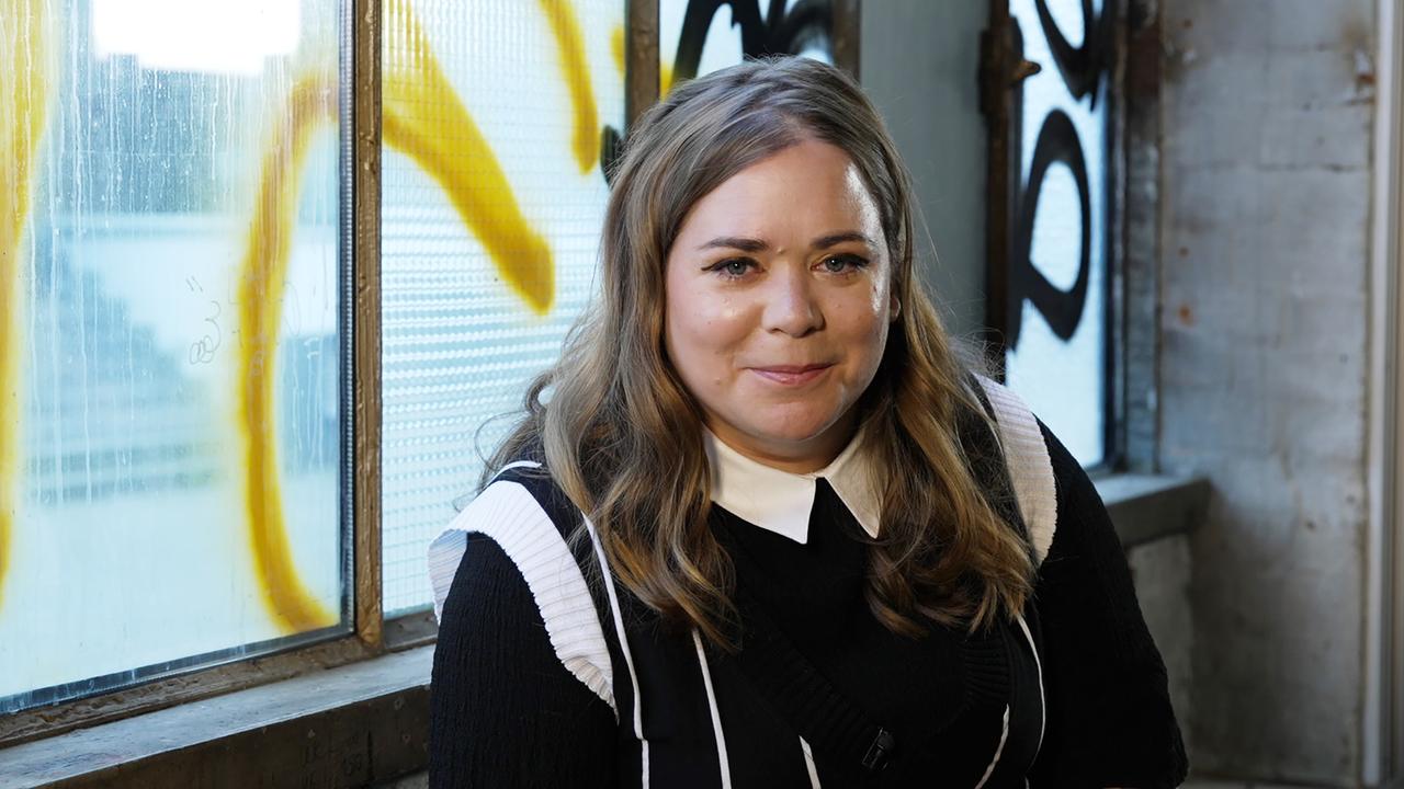 Stefanie Sargnagel vor einer mit Graffiti besprühten Glasscheibe.