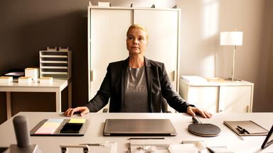 Ella (Annette Frier) sitzt an einem sehr ordentlichen Schreibtisch in einem Büro und hat beide Hände auf den Tisch gelegt. Vor ihr auf dem Tisch befinden sich Schreibwaren, ein Laptop, ein Handy und ein Tischtennisschläger, alles wohlgeordnet.