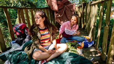 Jana (Zsa Zsa Hansen) und Nelly (Anais Cherif) sitzen nebeneinander in einem Baumhaus auf Schlafsäcken. Sie essen Ravioli aus der Dose.