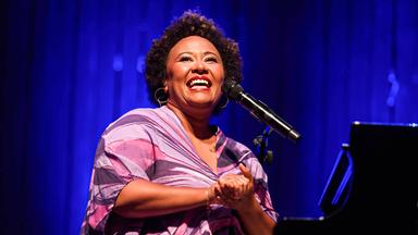 Eine Frau in einem bunten weiten Kleid klatscht in die Hände, während sie auf der Bühne an einem Mikro steht und lacht. Im Hintergrund ist ein Klavier zu sehen.