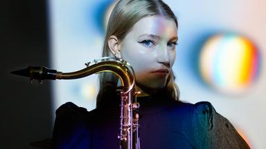 Die junge Musikerin Emma Rawicz, Kopf groß, seitlich. Das Sax seitlich vors Kinn haltend, der Saxbogen mit Mundstück gegen die Blickrichtung zeigend.