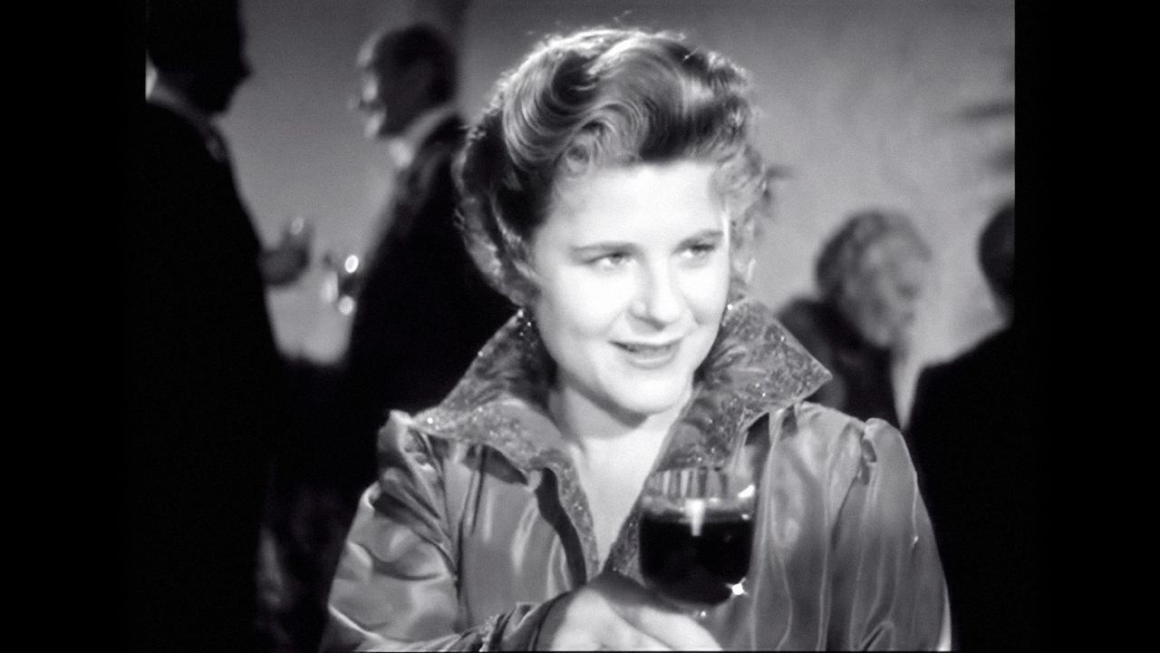 Das Bild zeigt eine Frau mit lockigem Haar, die ein Glas in der Hand hält. Sie trägt eine glänzende Jacke und lächelt. Im Hintergrund sind schemenhaft weitere Personen erkennbar.