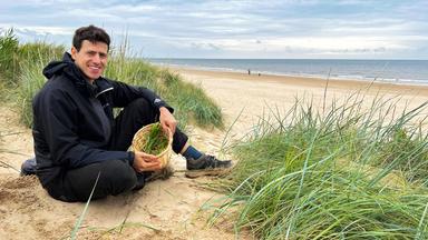 Ein Mann sitzt mit einem Korb mit Grüngewächs an einem Strand.