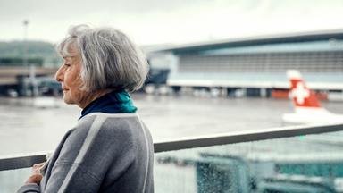Eine Frau steht am Flughafen Zürich und blickt in die Ferne. Im Hintergrund ist ein Teil des Flughafengebäudes und ein Flugzeug mit Schweizer Flagge zu sehen.