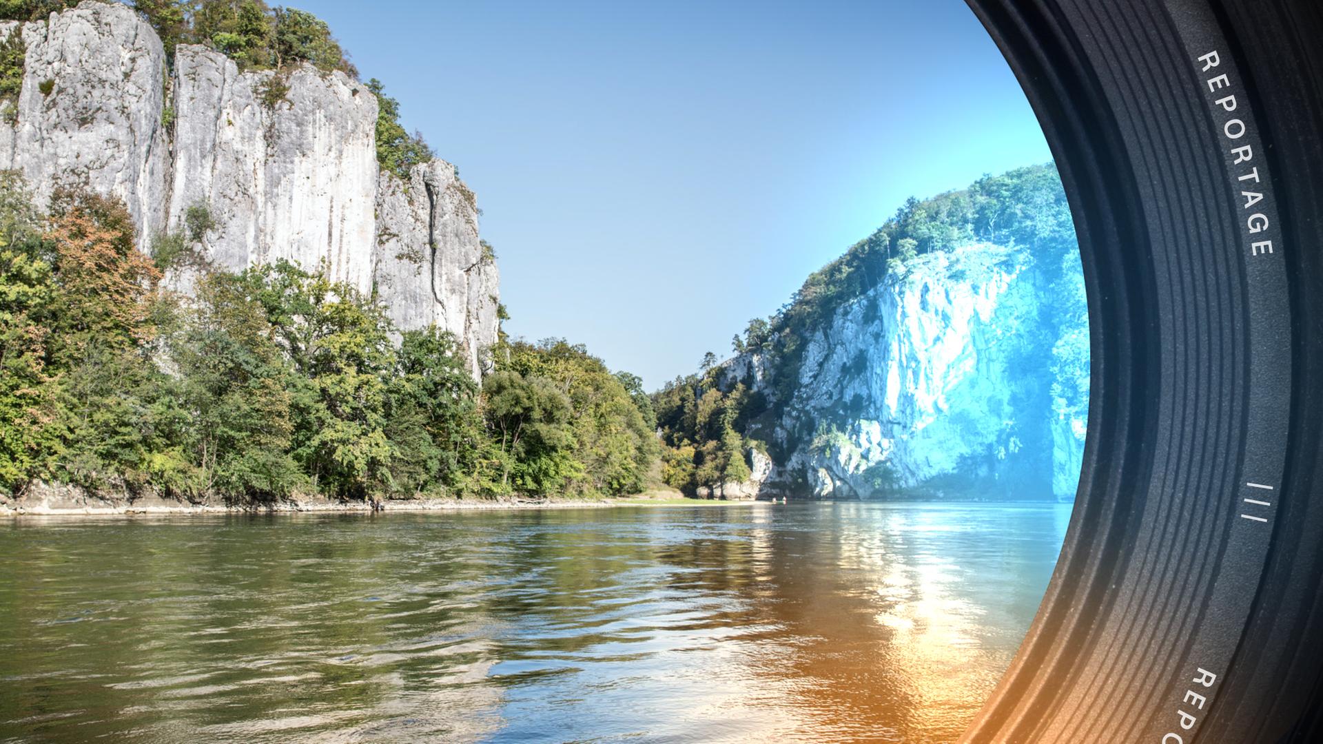Im Vordergrund klares Wasser. Im Hintergrund hohe Felsen, am Ufer Sträucher und Bäume.
