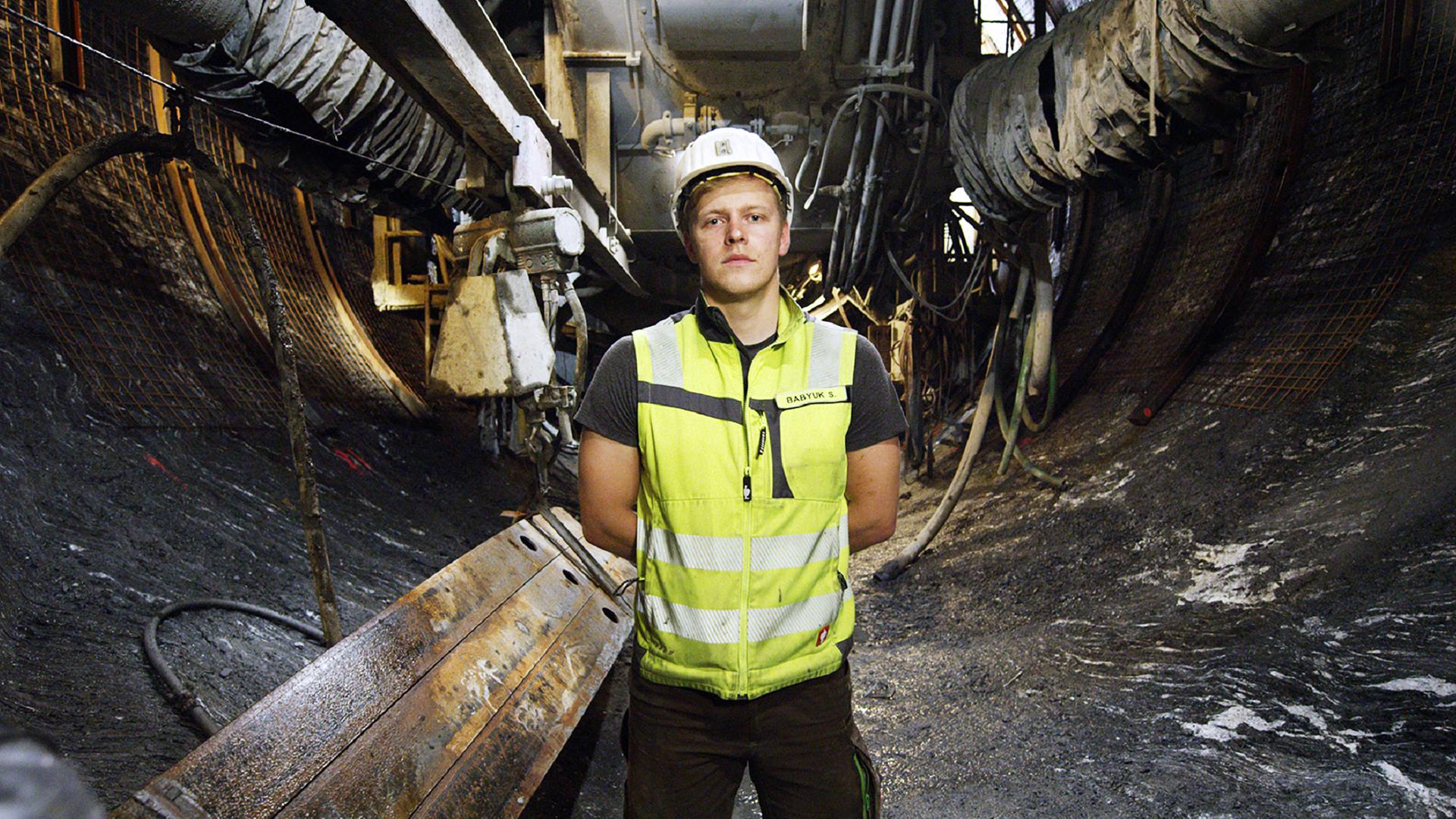 Ein junger Mann mit gelber Weste und weißem Schutzhelm steht in einem Bergtunnel frontal zu Kamera. Im Hintergrund sieht man Teile einer großen Maschine, die den Tunnel gräbt.