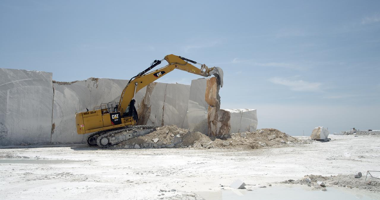Vor einem hohen hellbeigen Steinblock in einer Landschaft mit blauem Himmel und Sonnenschein steht ein gelber Bagger auf einem Steinhaufen und  bewegt mit dem Schaufelarm eine aus dem Block herausgeschnittene Marmorplatte.