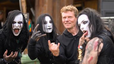 Ein Mann mit heller Haut und kurzen rötlichen Haaren steht zwischen vier weiß geschminkten Gestalten mit schwarzen langen Haaren. Alle machen typische Metal-Gesten.