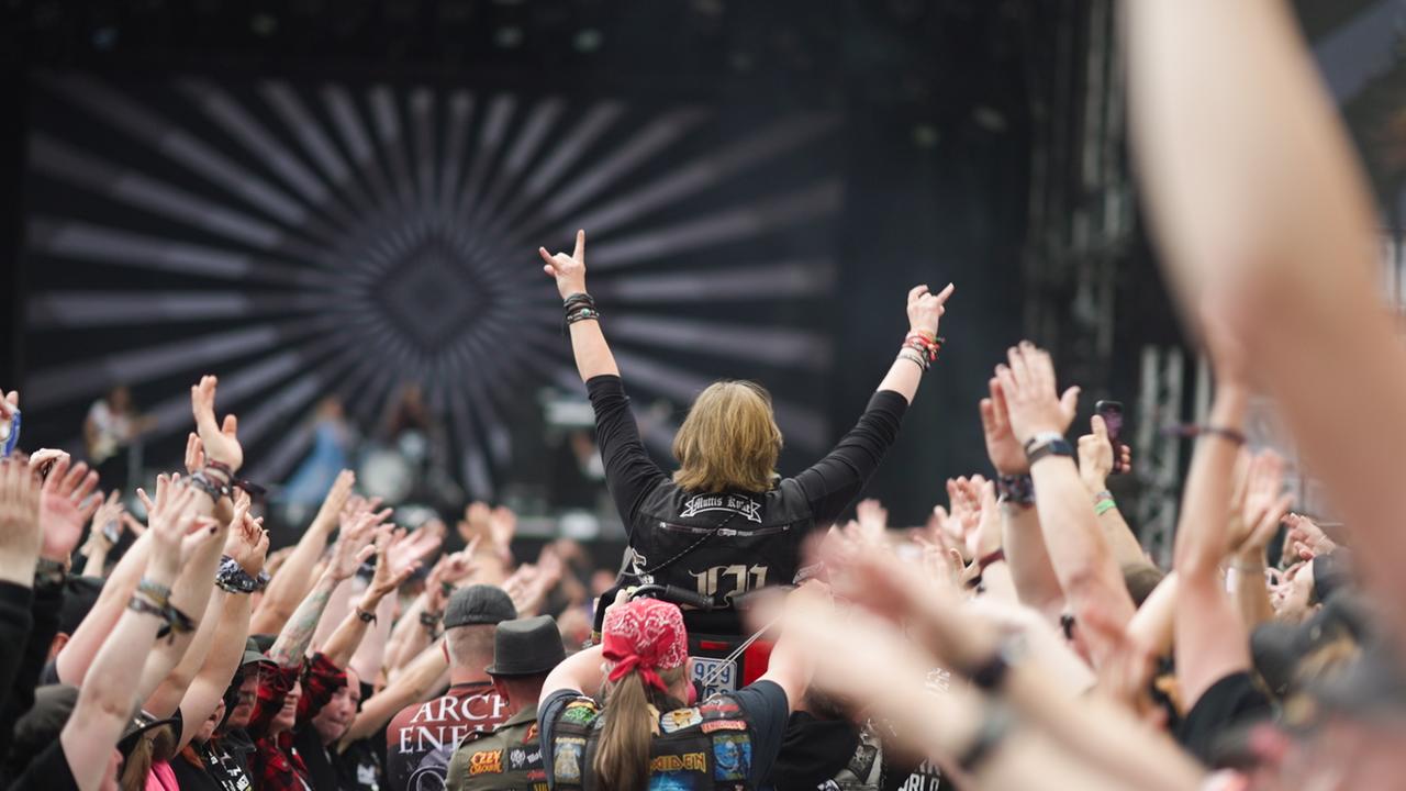 Eine Menge Menschen mit erhobenen Händen, in ihrer Mitte wird eine Frau getragen, sie hat beide Arme erhoben und macht den als „Pommesgabel“ bezeichneten Metal-Gruß.