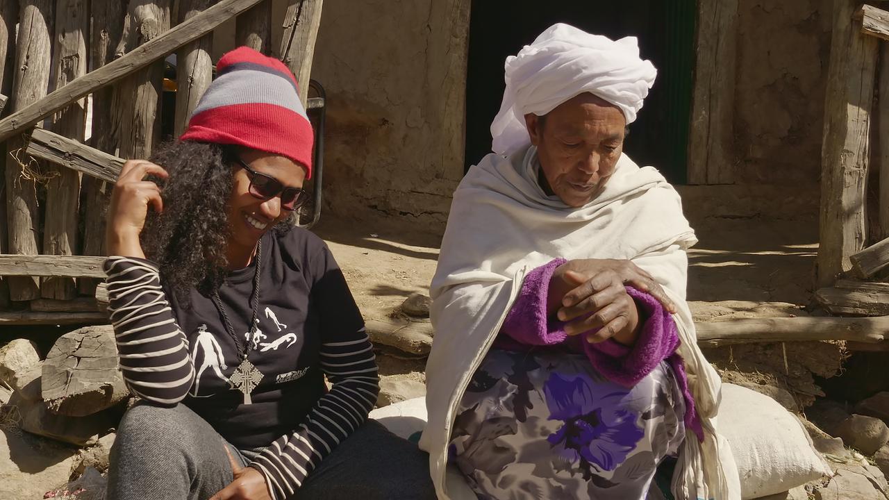 Links im Bild eine junge Frau mit Mütze und Brille, die lächelnd neben einer alten Frau in einem traditionellen Gewand sitzt.