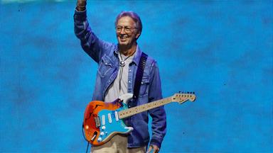 Eric Clapton steht mit einer Gitarre auf der Bühne und winkt.