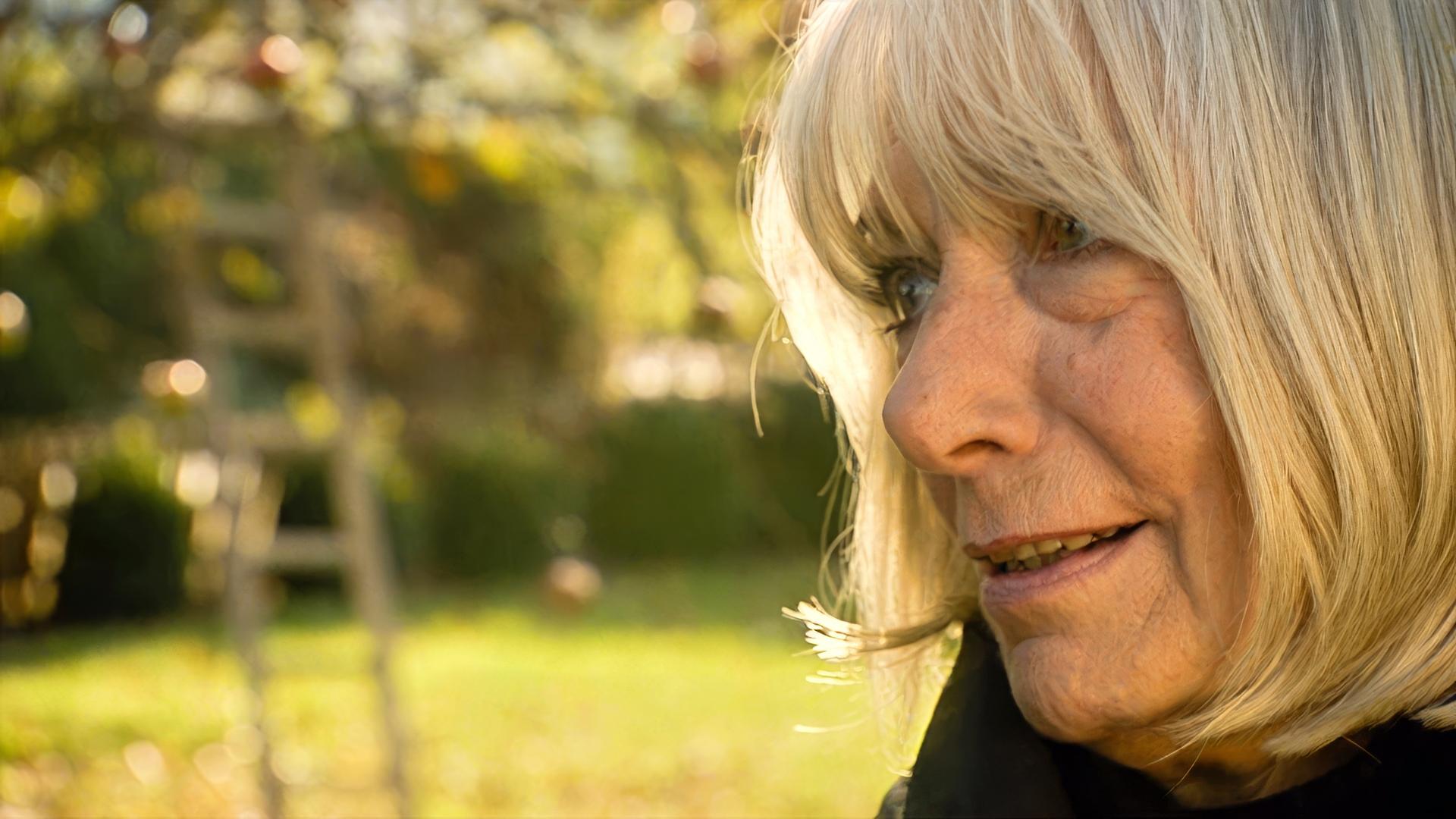 Das Bild zeigt eine Frau mit schulterlangen, blonden Haaren, die sanft ins Gesicht fällt. Sie hat einen freundlichen, nachdenklichen Ausdruck und trägt eine dunkle Jacke. Im Hintergrund ist eine unscharfe, grüne Landschaft mit einem Baum und einer unscharfen Leiter zu erkennen. Das Licht ist warm und hebt die Farben des Hintergrundes sanft hervor. Es scheint eine ruhige, naturnahe Umgebung zu sein, die eine gewisse Nostalgie vermittelt.