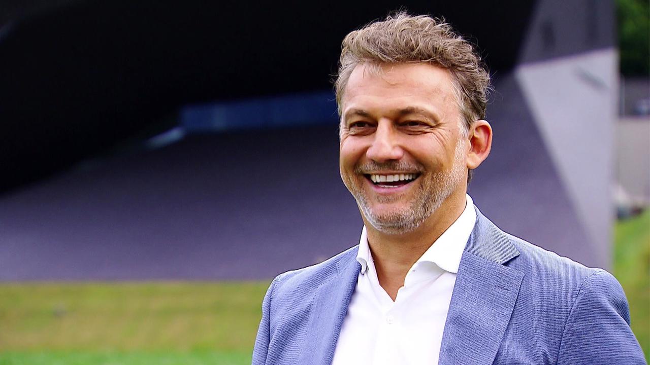 Das Bild zeigt einen Mann, der freundlich lächelt. Er trägt einen hellen Anzug und ein weißes Hemd. Im Hintergrund ist ein modernes, architektonisch gestaltetes Gebäude zu sehen, das zu den Tiroler Festspielen in Erl gehört. Die Landschaft ist grün, mit einer Wiese, die sich bis zum Gebäude erstreckt. Der Himmel ist klar und das Licht vermittelt eine freundliche, sonnige Atmosphäre. Diese Szenerie spiegelt die kulturellen Aktivitäten und die besondere Umgebung der Tiroler Festspiele wider.