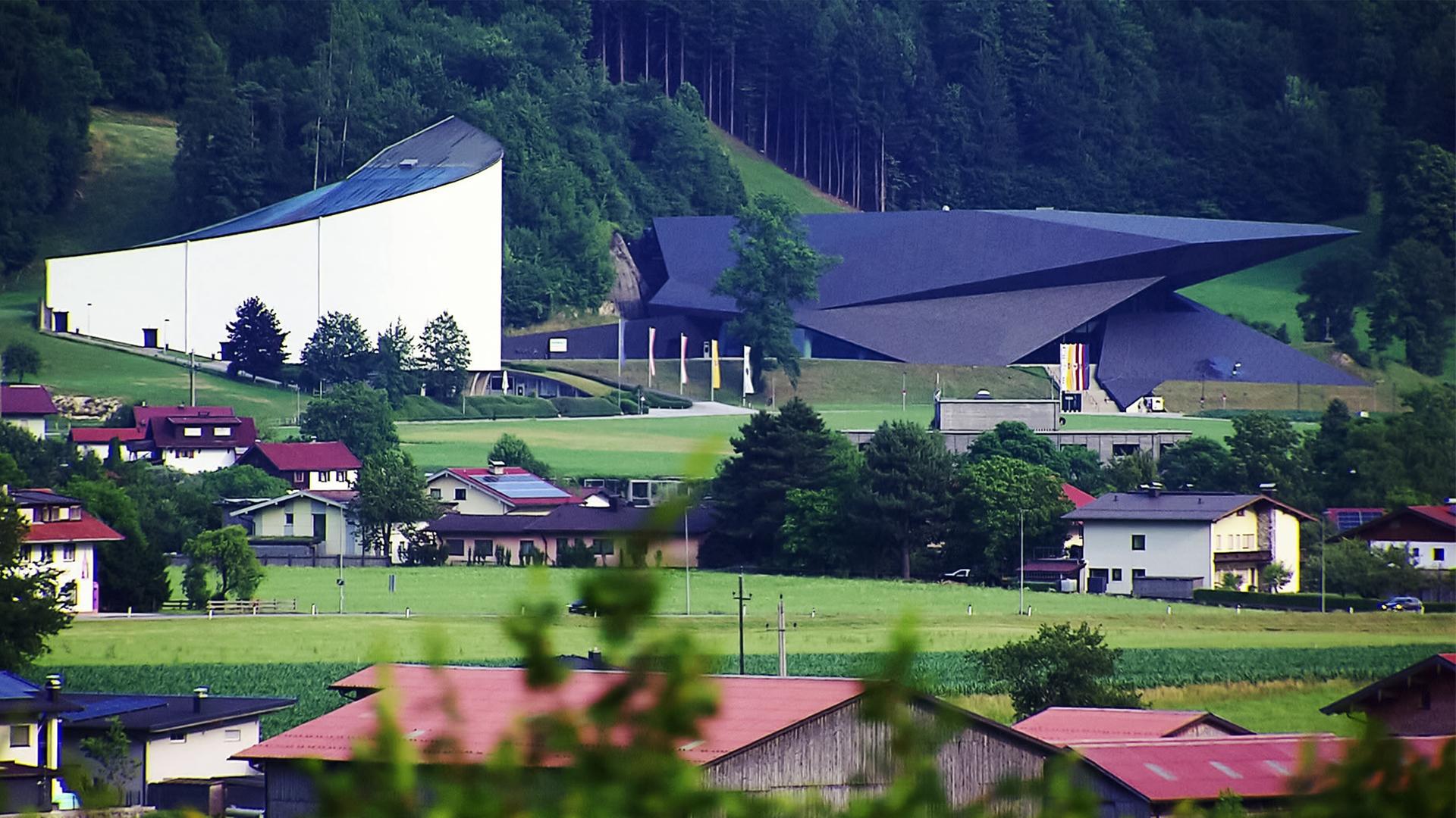 Das Bild zeigt das Festspielhaus Erl, ein markantes architektonisches Gebäude, das in eine grüne Landschaft integriert ist. Es befindet sich im Tiroler Dorf Erl, nahe der bayerischen Grenze. Auf der linken Seite sieht man die hellere, geschwungene Fassade des Festspielhauses, die sich harmonisch in die Umgebung einfügt. Daneben befindet sich ein dunkleres Gebäude mit einer ausgefallenen, spitzen Dachform.   Im Vordergrund sind mehrere Wohnhäuser mit roten Dächern und Gärten zu erkennen, die in einer hügeligen Landschaft angeordnet sind. Die Umgebung ist geprägt von grünen Wiesen, Bäumen und sanften Hügeln, die die Idylle des ländlichen Bereichs unterstreichen. Im Hintergrund sind dichte Wälder zu sehen, die sich bis zu den Gipfeln der Berge hochziehen.   Das Bild vermittelt einen Eindruck von der ruhigen Atmosphäre und der kulturellen Bedeutung der Tiroler Festspiele, die in dieser malerischen Umgebung stattfinden.