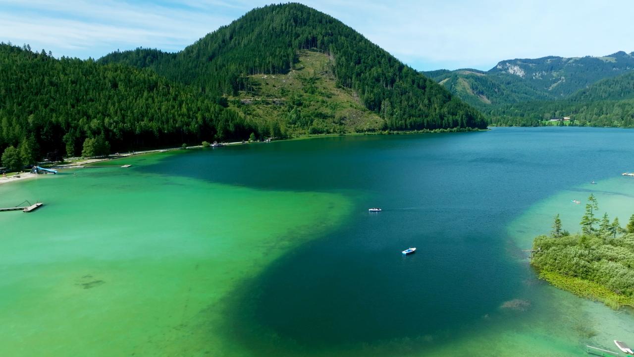 Das Bild zeigt eine beeindruckende Landschaft im Erlauftal. Im Vordergrund befindet sich der Erlaufsee, dessen Wasser in verschiedenen Blau- und Grüntönen schimmert. Die Wasseroberfläche ist ruhig und spiegelt die umgebenden Berge wider.   Rund um den See erheben sich grüne Hügel und steile, bewaldete Berge, die von Nadelbäumen bewachsen sind. Ein pfadförmiger Strandbereich mit hellerer Sandfarbe ist an einer Seite des Sees sichtbar, wo einige kleine Boote auf dem Wasser liegen. Im Hintergrund sind weitere Berge und Waldflächen zu sehen, die die alpine Umgebung betonen.  Die Atmosphäre des Bildes strahlt eine friedliche und naturnahe Umgebung aus, die das typische Landschaftsbild des Mostviertels widerspiegelt.