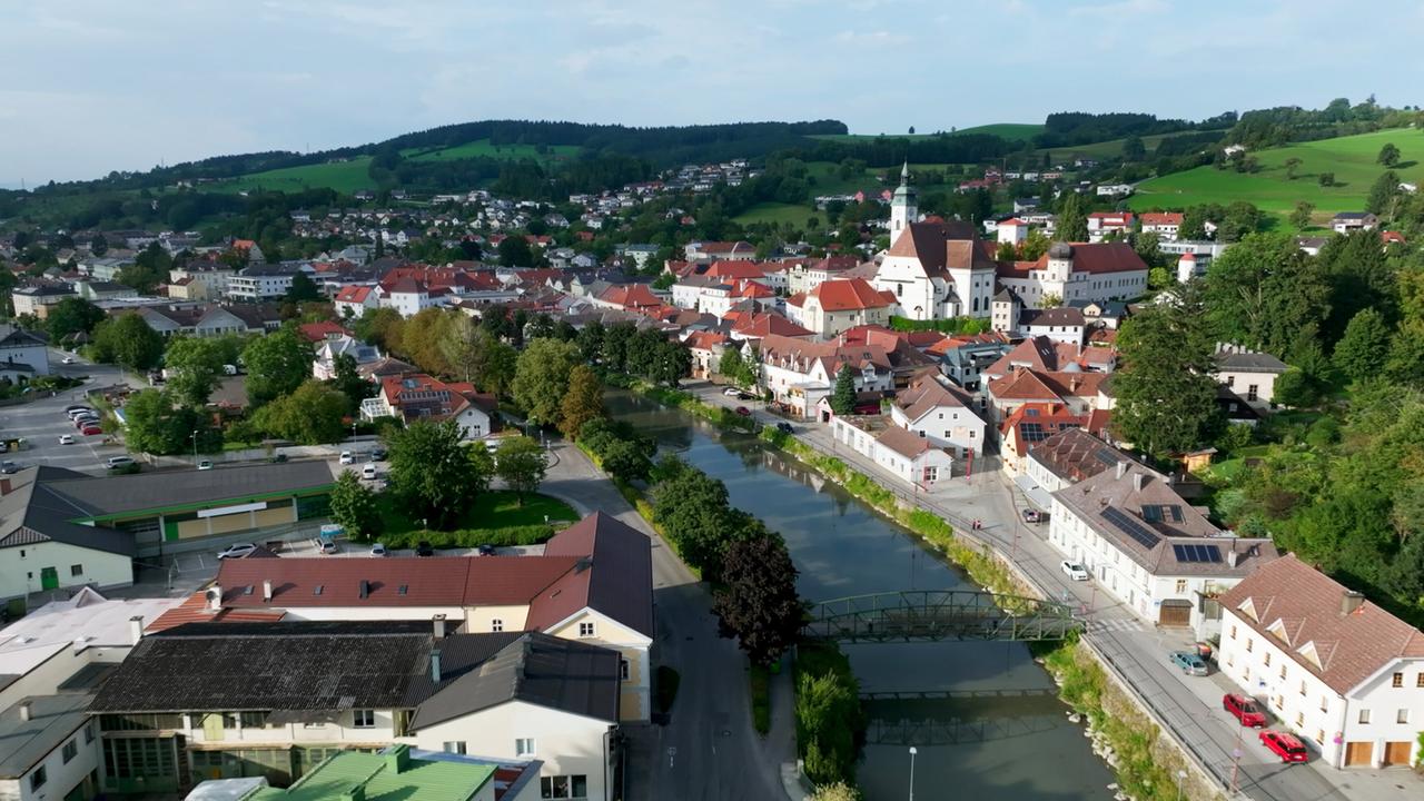 Das Bild zeigt eine Luftaufnahme der Stadt Scheibbs im Erlauftal, umgeben von einer beeindruckenden Landschaft. Im Vordergrund fließt ein schmaler Fluss, der von grünen Ufern gesäumt ist. Auf der linken Seite sind verschiedene Gebäude zu sehen, darunter Einfamilienhäuser mit roten Dächern und einige größere Bauwerke. Im Hintergrund erheben sich sanfte Hügel, die mit Bäumen und Wiesen bedeckt sind. Die Architektur der Stadt ist traditionell mit einer Mischung aus älteren und neueren Gebäuden. Die Atmosphäre wirkt ruhig und ländlich, unterstrichen von der klaren Natur und den historisch anmutenden Strukturen.