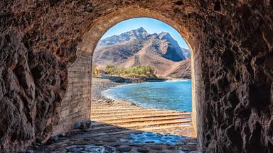 Ansicht eines Steintunnels, der zu einem Strand vor Bergen führt.