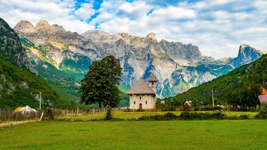 Eine kleine kastige Steinkirche vor dem Hintergrund eines Alpenpanoramas.