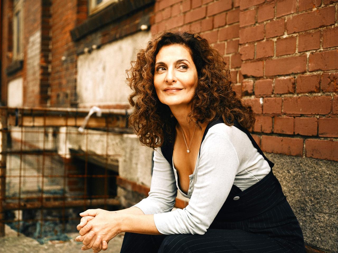 Das Bild zeigt eine Frau mit lockigem, braunem Haar, die auf einer Mauer sitzt. Sie trägt ein graues Oberteil und eine schwarze Latzhose mit weißen Streifen. Ihre Arme sind auf den Knien abgelegt, und sie hat ein entspanntes Lächeln auf dem Gesicht. Der Hintergrund besteht aus einer Ziegelwand und einer unvollendeten, verwitterten Wandstruktur, die auf eine Baustelle hinweist. Im Bild sind zudem metallische Bauelemente sichtbar, die an einen Bauzaun erinnern. Die Lichtverhältnisse sind warm, was eine freundliche Atmosphäre erzeugt.