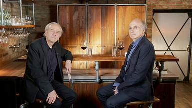 Das Bild zeigt zwei Männer, die an einer Bartheke sitzen. Der Raum hat eine rustikale Atmosphäre mit Backsteinmauern und Holzelementen. Die Bartheke ist aus Holz und Metallrohren gefertigt. Auf der Theke stehen Gläser, darunter ein Glas mit rotem Wein und ein weiteres mit Wasser.   Der Mann links trägt einen schwarzen Anzug mit einem schwarzen Hemd und hat graues Haar. Er blickt direkt in die Kamera, wirkt nachdenklich. Der Mann rechts sitzt in einem dunklen, strukturierten Blazer und einem blauen Hemd. Er hat kurze, blonde Haare und trägt eine Brille. Auch er schaut in die Kamera, hat einen ernsten Gesichtsausdruck.   Im Hintergrund sind weitere Regale mit Gläsern zu sehen, und an der Wand hängen Lampen, die für indirektes Licht sorgen. In der Ecke ist eine Tür zu erkennen, die einen schlichten Design hat. Die Atmosphäre des Raumes wirkt freundlich und einladend, geeignet für Gespräche oder Interviews.