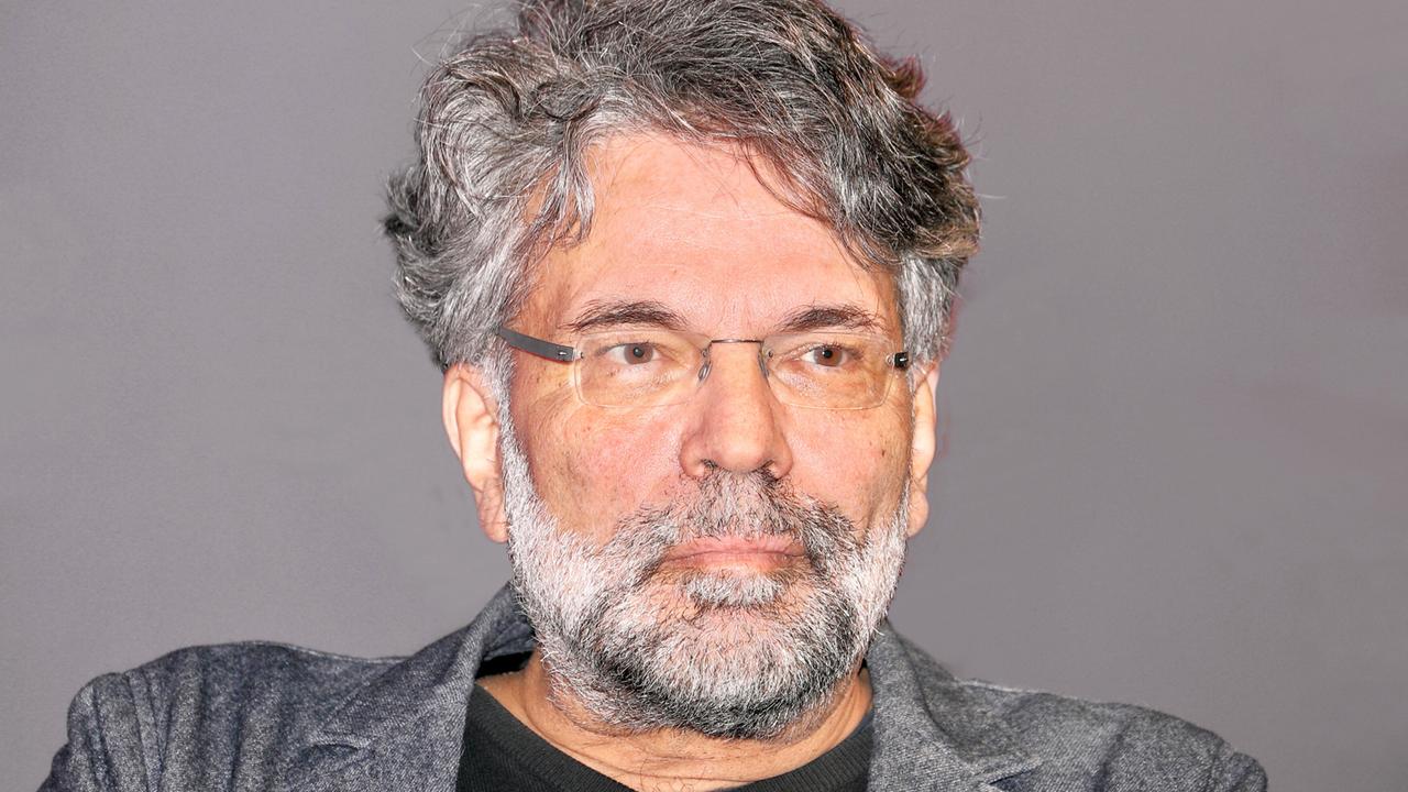 Das Bild zeigt einen Mann mittleren Alters mit grauen, leicht gewellten Haaren und einem dichten, grauen Bart. Er trägt eine dunkle, lässige Oberbekleidung sowie ein schwarzes T-Shirt. Sein Blick ist ernst und nachdenklich, während er in die Kamera schaut. Im Hintergrund ist eine einfarbige, schlichte graue Wand zu sehen. Der Fokus des Bildes liegt auf dem Gesicht des Mannes, das von einer Brille und einem intelligenten Ausdruck geprägt ist. Die Gesamtatmosphäre vermittelt einen Eindruck von Ernsthaftigkeit und Intellektualität.