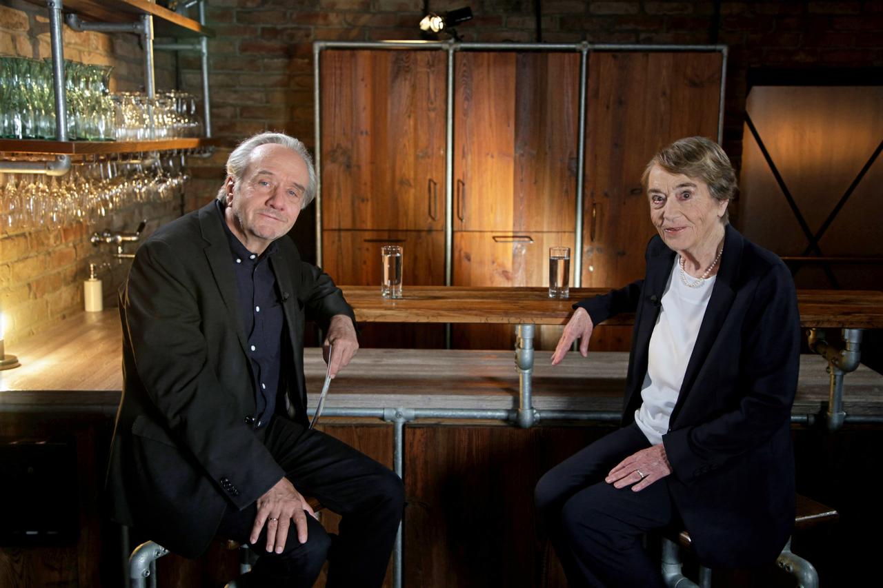 Das Bild zeigt zwei Personen, die an einer hölzernen Theke sitzen. Die Theke ist aus einem dunklen Holz gefertigt und hat metallische Beine. Im Hintergrund sind Regale mit Gläsern zu sehen, die an einer Ziegelwand angebracht sind.   Eine Person ist ein Mann mit grauen, kurzen Haaren und trägt einen dunklen Anzug. Er sitzt auf einem Barstuhl, lächelt leicht und sieht direkt in die Kamera. Die andere Person ist eine Frau mit kurzen, blonden Haaren, die einen schlichten, dunklen Blazer und ein weißes Oberteil trägt. Sie sitzt ebenfalls auf einem Barstuhl und hat die linke Hand auf der Theke abgelegt. Vor ihr steht ein Glas mit Wasser.  Die gesamte Szene hat eine warme, einladende Atmosphäre und ist durch gezielte Beleuchtung gut ausgeleuchtet. Der Hintergrund ist eher neutral gehalten, sodass die beiden Personen im Vordergrund hervorgehoben werden.