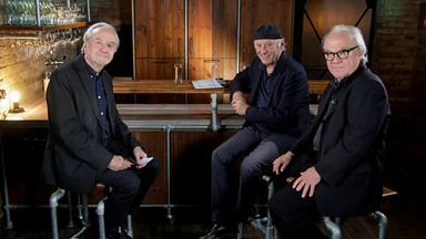 Das Bild zeigt drei Männer, die in einem gemütlich gestalteten Innenraum sitzen. Der Hintergrund besteht aus einer Steinwand, die mit Holzregalen dekoriert ist, auf denen Gläser stehen. In der Mitte des Bildes verläuft eine Bar aus Holz, hinter der sich eine geschlossene Holzschranktür befindet.   Die drei Männer sind auf Barhockern platziert. Der Mann links hat graue Haare, trägt einen dunklen Anzug und schaut freundlich in die Kamera. Der Mann in der Mitte trägt ein schwarzes Hemd und eine schwarze Kappe, lächelt und hat seine Hände auf der Tischkante abgelegt. Der Mann rechts trägt eine dunkle Brille und eine schwarze Jacke. Er sitzt mit verschränkten Armen, hat einen ernsten, aber freundlichen Gesichtsausdruck.  Die Beleuchtung ist warm und sorgt für eine einladende Atmosphäre. Insgesamt vermittelt das Bild den Eindruck eines informellen Gesprächs zwischen den drei Männern.