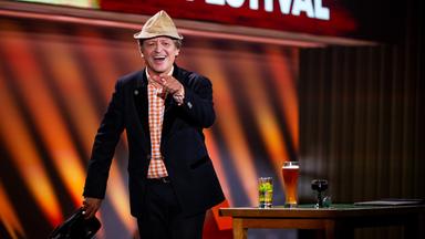 Erwin Pelzig steht in der Mitte des Bildes und deutet mit seiner linken Hand in Richtung Kamera. Er trägt einen Hut. Im Hintergrund steht auf der rechten Seite der Bühne ein Tisch.