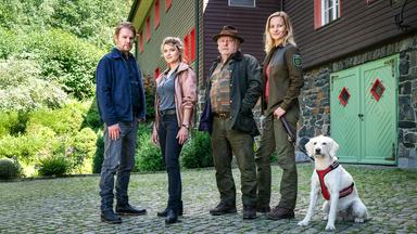 Vier Personen und ein weißer Hund stehen vor einem Forsthaus. Die Personen tragen Outdoor-Kleidung, der Hund trägt ein Geschirr.