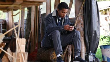Ado Nyangabo (Seedy Touray) sitzt auf einem Holzhocker in einer Werkstatt und schaut auf sein Handy.