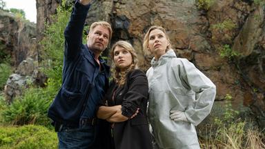 Vor einer Felswand stehend zeigt Kommissar Winkler (Kai Scheve) Kommissarin Karina Szabo (Lara Mandoki) und Pathologin Dr. Elena Kulikova (Masha Tokareva) die Absturzstelle.