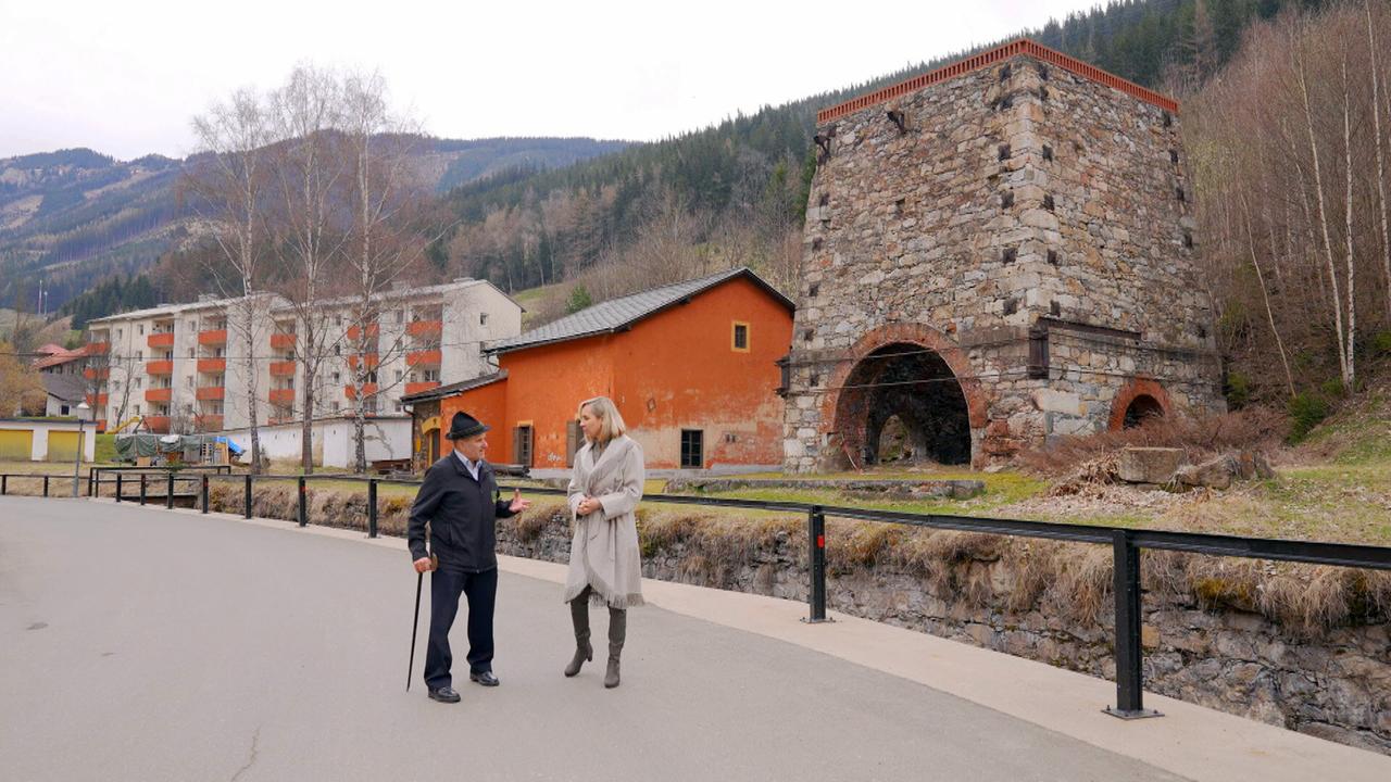 Das Bild zeigt eine Szene, die in der Stadt Vordernberg, Österreich, aufgenommen wurde. Im Vordergrund stehen zwei Personen: ein älterer Mann mit einem Stock und eine Frau. Beide sprechen miteinander. Der Mann trägt einen schwarzen Hut und eine schwarze Jacke, während die Frau einen beigen Mantel und graue Stiefel trägt.  Im Hintergrund sind mehrere Gebäude zu sehen, darunter ein mehrstöckiges grau-weißes Gebäude mit orangefarbenen Balkonen. Zu anderen Gebäuden gehören ein orangefarbener Bau und ein kleinerer, weißer Anbau, die in der Nähe eines alten Steingebäudes stehen.  Das Steingebäude hat einen runden Durchgang an der Unterseite und ist teilweise verfallen. Um das Gebäude herum gibt es einige kahle Stellen mit wenig Vegetation, und im Hintergrund erheben sich bewaldete Hügel. Der Himmel ist bewölkt, und die Umgebung vermittelt den Eindruck eines ruhigen, industriellen Erbes.