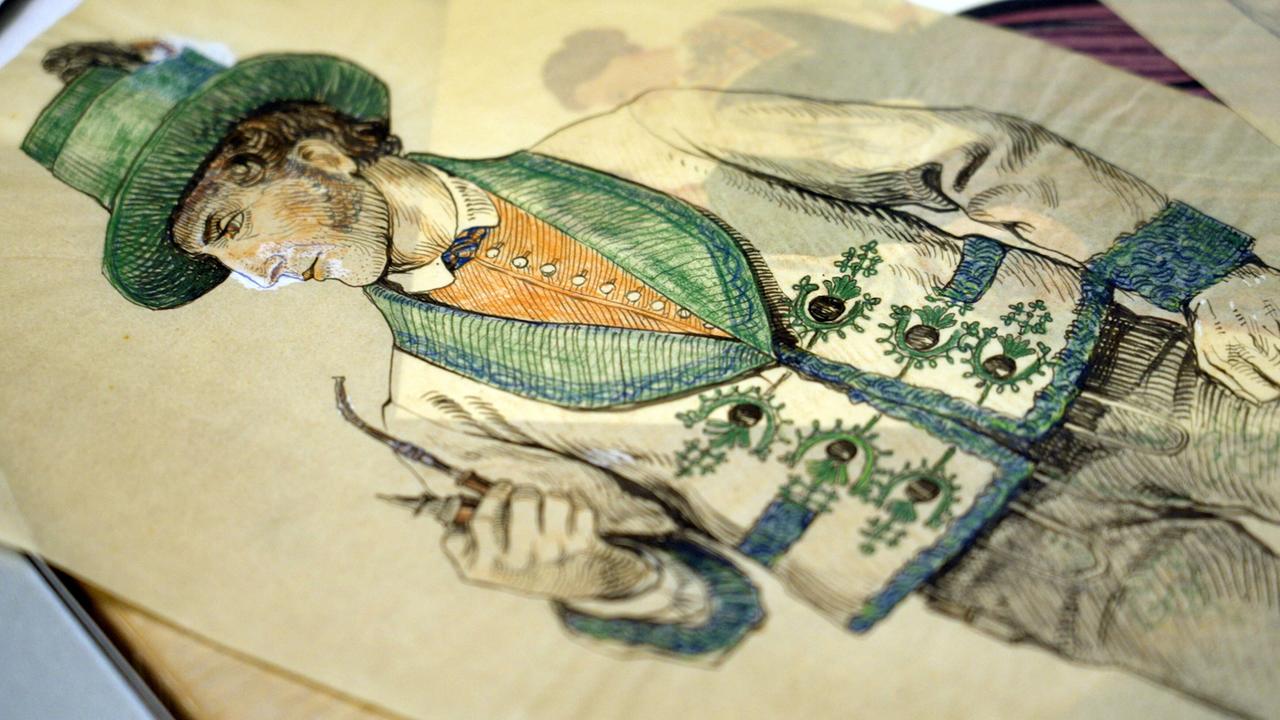 Das Bild zeigt eine historische Zeichnung eines Mannes in traditioneller Tracht. Der Mann trägt einen grünen Hut und ist in eine Jacke mit aufwendigen Verzierungen gekleidet. Die Jacke ist in hellen Farben, mit grünen und orangen Elementen, sowie kunstvollen Stickereien geschmückt. Der Mann hält in einer Hand ein kleines, braunes Objekt, möglicherweise einen Stock oder ein Werkzeug. Der Hintergrund des Bildes ist unscharf, und es sind auch andere Zeichnungen und Papiere sichtbar, die auf einem Tisch oder einer anderen Oberfläche liegen. Das Bild strahlt ein Gefühl von Tradition und historischem Erbe aus.