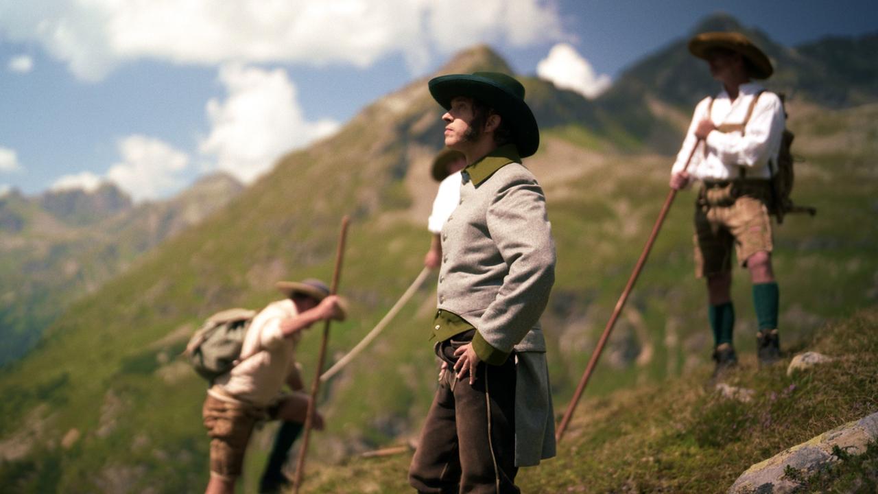 Die Abbildung zeigt mehrere Männer in traditioneller Bergsteigerkleidung, die auf einem Hügel in einer alpinen Landschaft stehen. Der Fokus liegt auf einem Mann in der Mitte, der einen grauen Anzug mit grünen Akzenten trägt und einen breiten, dunklen Hut auf hat. Er blickt nach oben in die Ferne.   Im Hintergrund sieht man die majestätischen Berge der Schladminger Tauern, umgeben von grünem Gras und felsigen Abhängen. Ein weiterer Mann steht leicht im Schatten rechts hinter dem Hauptmann, während ein dritter Mann sich etwas weiter links mit einem Wanderstock auf einen Stein stützt.   Der Himmel ist blau mit vereinzelten weißen Wolken. Die Szenerie vermittelt ein Gefühl von Abenteuer und der Erkundung der Natur.