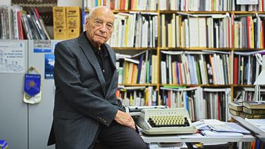 Das Porträtfoto des Dichters Eugen Gomringer zeigt einen älteren Herr im dunklen Anzug, der in einem Büro mit Bücherwänden halb auf einem Schreibtisch sitzt, auf dem eine mechanische Schreibmaschine steht.