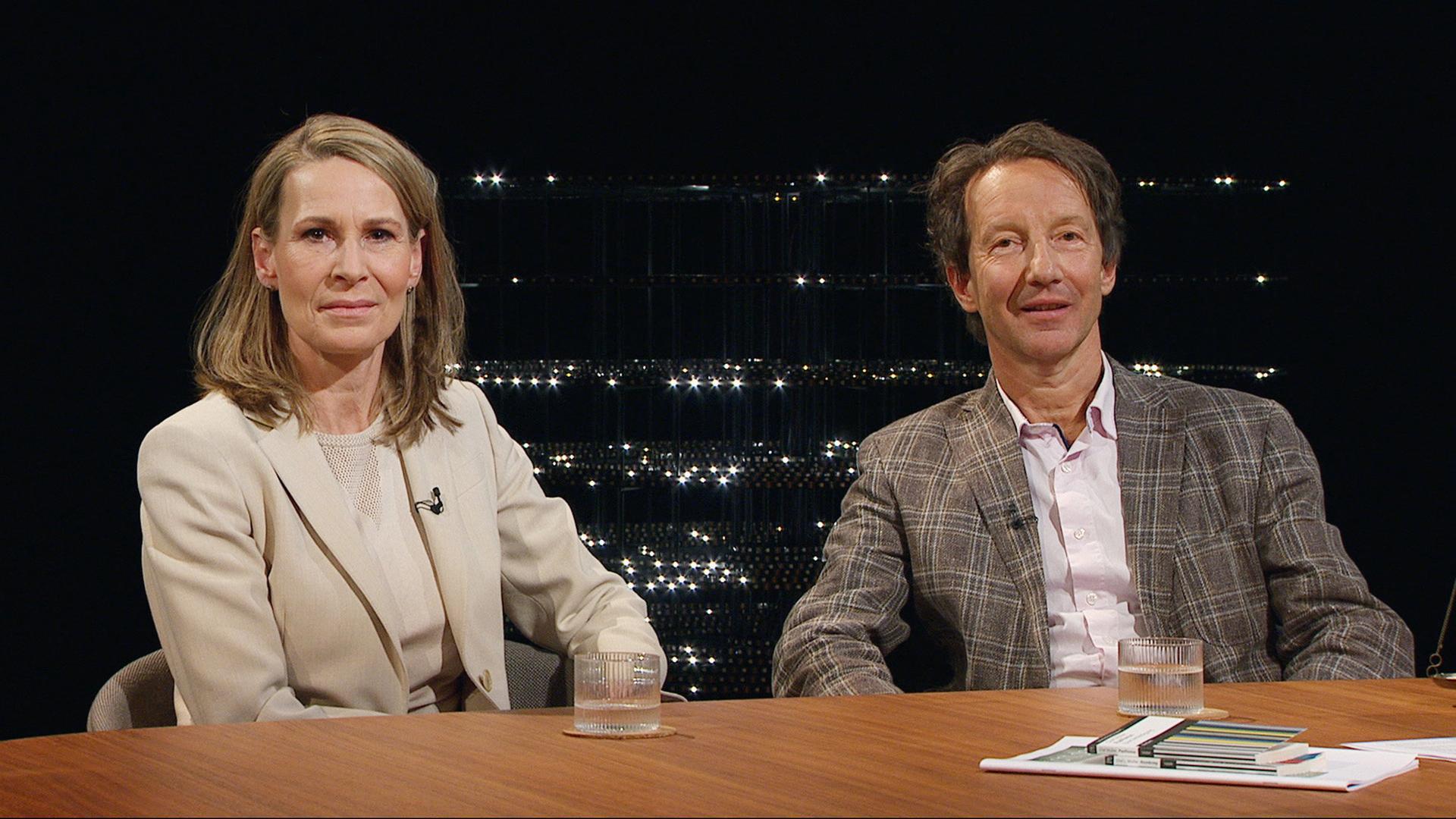 Katja Gentinetta und Olaf Müller sitzen bei Sternstunde Philosophie an einem Studiotisch mit schwarzen Hintergrund und Lichtinstallation, beide tragen helle Jackets.