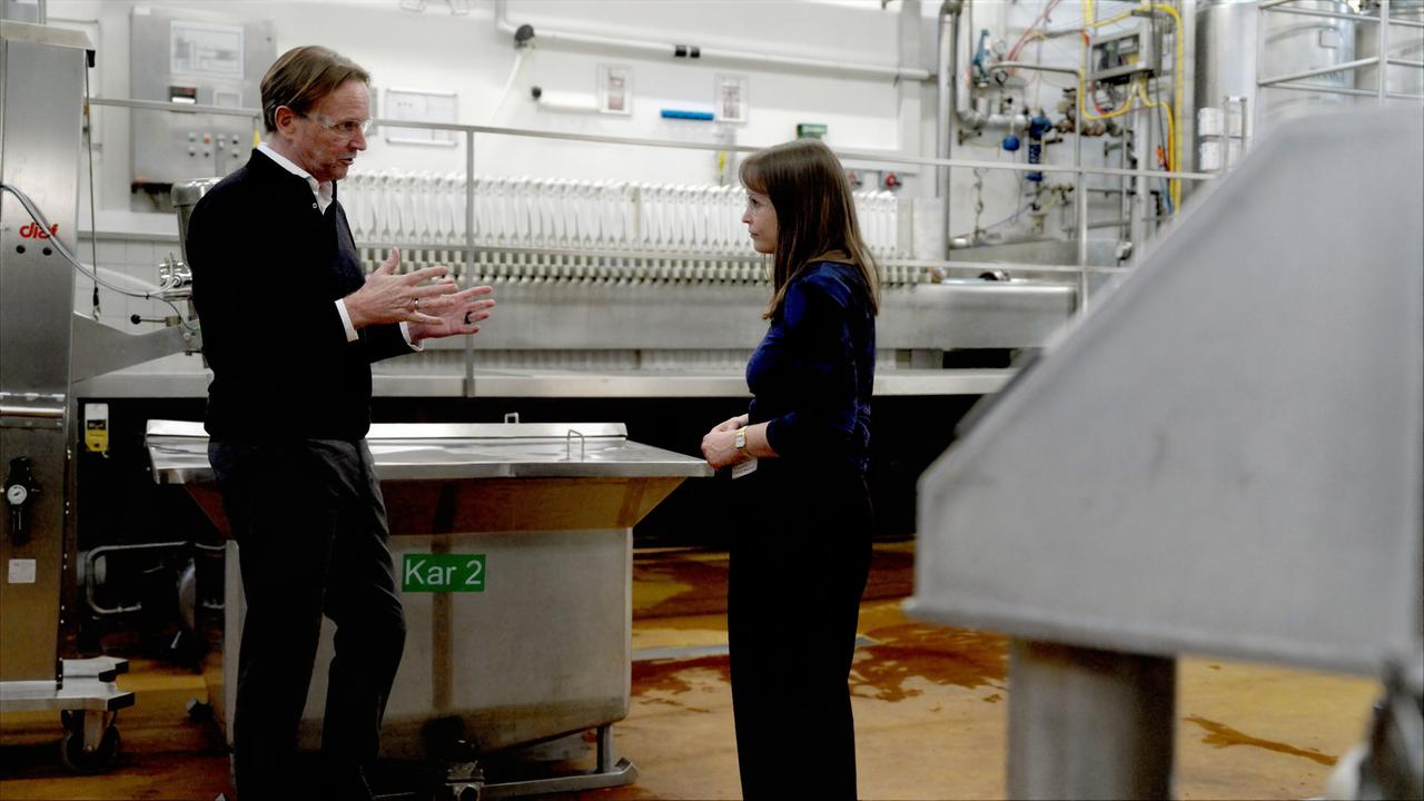 Das Bild zeigt eine Gesprächsszene in einem modernen, industriellen Innenraum. Auf der linken Seite steht ein älterer Mann mit kurzen, grauen Haaren, der in einem schwarzen Pullover und einer weißen Bluse spricht. Er verwendet seine Hände zur Betonung, während er seine Argumente vorträgt. Auf der rechten Seite steht eine jüngere Frau mit langen, braunen Haaren, die in einem blauen Oberteil gekleidet ist. Sie blickt aufmerksam auf den Mann und hat eine Hand leicht erhoben, möglicherweise in einer Fragestellung oder Reaktion.  Im Hintergrund sieht man verschiedene industrielle Geräte aus Edelstahl, darunter große, funktionale Tanks und Ausrüstungen. An den Wänden sind mehrere Schaltschränke mit Tasten und Anzeigen zu erkennen. Der Boden ist aus einem braunen Material und weist einige Flecken auf, was auf eine industrielle Umgebung hinweist. Der Gesamteindruck ist sachlich und informativ, passend zur Thematik der Diskussion über pharmazeutische Herausforderungen und technologische Abhängigkeiten in Europa.