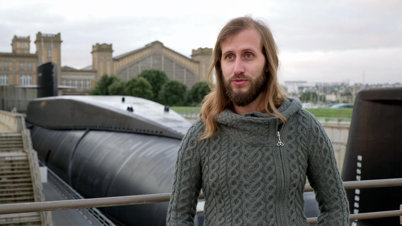 Das Bild zeigt einen Mann mit langen, lockigen, blonden Haaren und einem Vollbart, der einen grauen, grob gestrickten Pullover trägt. Er steht an einem Geländer und blickt in die Kamera. Hinter ihm ist ein großes, schwarzes U-Boot zu sehen, das im Hafen liegt. Im Hintergrund erkennt man ein historisches Gebäude mit einer großen Glasfassade und mehreren Türmen, das eine industrielle Architektur aufweist. Der Himmel ist bewölkt und es herrscht eine graue Lichtstimmung. Das Bild vermittelt eine Atmosphäre von Technik und maritimer Umgebung.