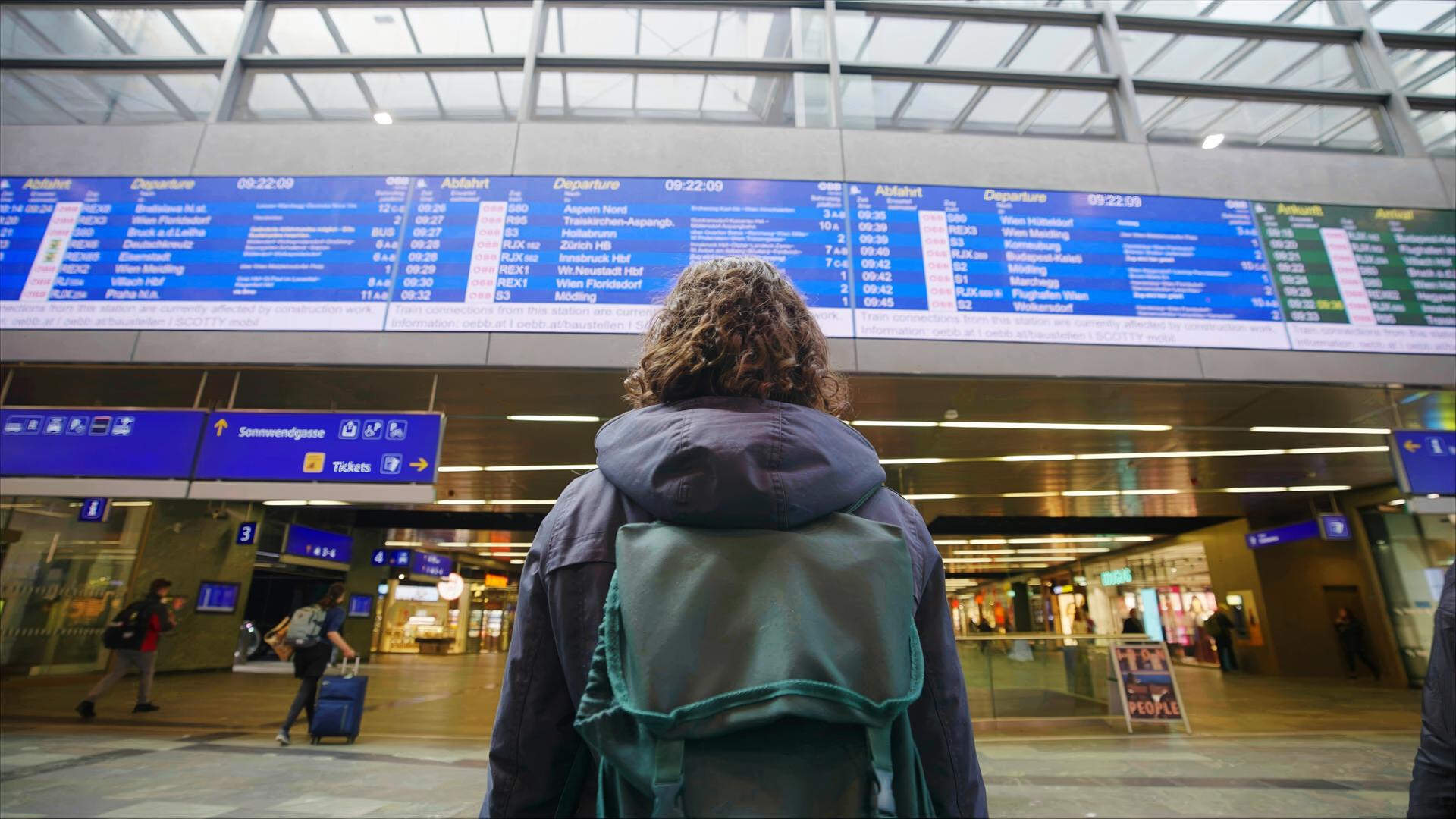 Das Bild zeigt eine Person mit lockigem Haar, die in einer modernen, hellen Bahnhofshalle steht. Die Person trägt eine grüne Rucksacktasche und schaut auf eine große Anzeigetafel über ihr. Diese Tafel zeigt die Abfahrtszeiten von Zügen mit Zielen, die teilweise in deutscher Sprache vermerkt sind. Die Halle hat große Fenster, durch die Tageslicht strömt, und eine offene, zugängliche Architektur. Auf dem Boden sind andere Reisende zu sehen, die mit Gepäck in verschiedenen Richtungen gehen. An den Wänden sind Informationsschilder, und es gibt einen Bereich mit Ticketautomaten. Insgesamt vermittelt die Umgebung ein geschäftiges Reisewohlgefühl.