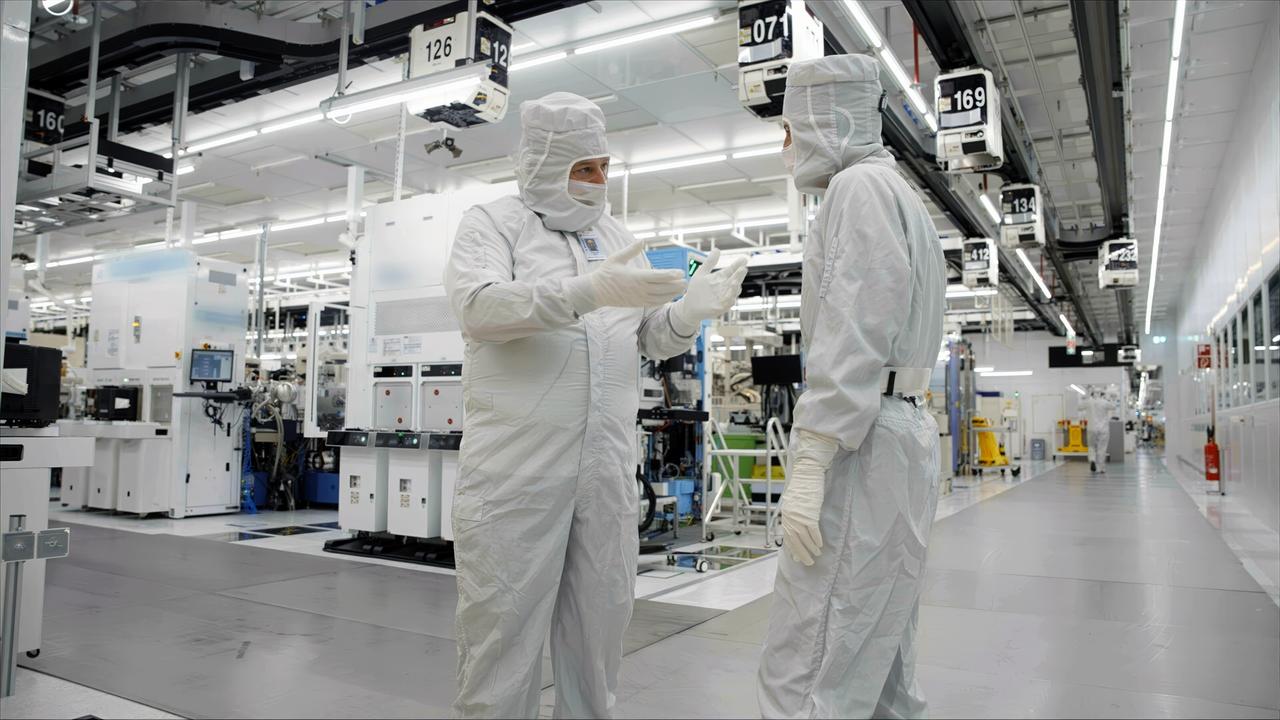 Das Bild zeigt einen Reinraum in einer Mikrochip-Produktionsanlage. Zwei Personen sind im Vordergrund zu sehen, die in spezieller Schutzkleidung gekleidet sind, bestehend aus weißen Overalls, Hauben und Handschuhen. Sie stehen sich gegenüber und scheinen sich zu unterhalten, während einer von ihnen ein kleines, flaches Objekt in der Hand hält.   Im Hintergrund erstrecken sich zahlreiche Maschinen und Geräte, die für die Mikrochip-Produktion verwendet werden. Die Wände sind in einem hellen Weiß gehalten und die Beleuchtung ist intensiv, was eine sterile Umgebung schafft. An den Wänden hängen Nummernschilder, die verschiedene Bereiche der Produktionsstätte kennzeichnen. Der Boden ist glatt und ebenfalls hell gefärbt, was den Eindruck von Sauberkeit und Ordnung verstärkt.   Diese Umgebung ist typisch für Hochtechnologieanlagen, in denen präzise und empfindliche Arbeiten durchgeführt werden.