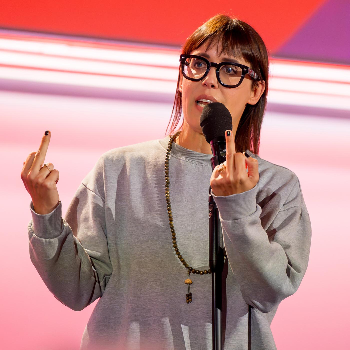 Eva Karl Faltermeier trägt einen grauen Pullover. Sie zeigt beide Mittelfinger. Sie trägt eine Brille. Der Hintergrund ist hell und rot beleuchtet.