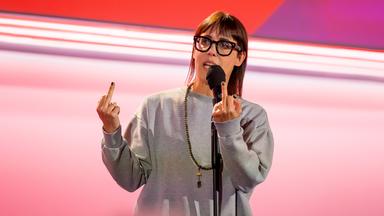 Eva Karl Faltermeier trägt einen grauen Pullover. Sie zeigt beide Mittelfinger. Sie trägt eine Brille. Der Hintergrund ist hell und rot beleuchtet.