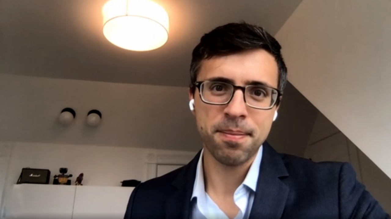 Ein Mann mit Brille und Anzug sitzt mit Kopfhörern vor dem Hintergrund eines weißen Schrankes und einer Lampe