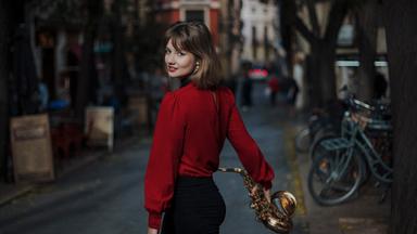 Die junge Saxophonistin Fabia Mantwill in roter Bluse und schwarzer Hose, von der Kamera auf der Straße in einer Wohngegend weglaufend, sich umdrehend in der Bewegung und das Saxophon in der rechten Hand habend.